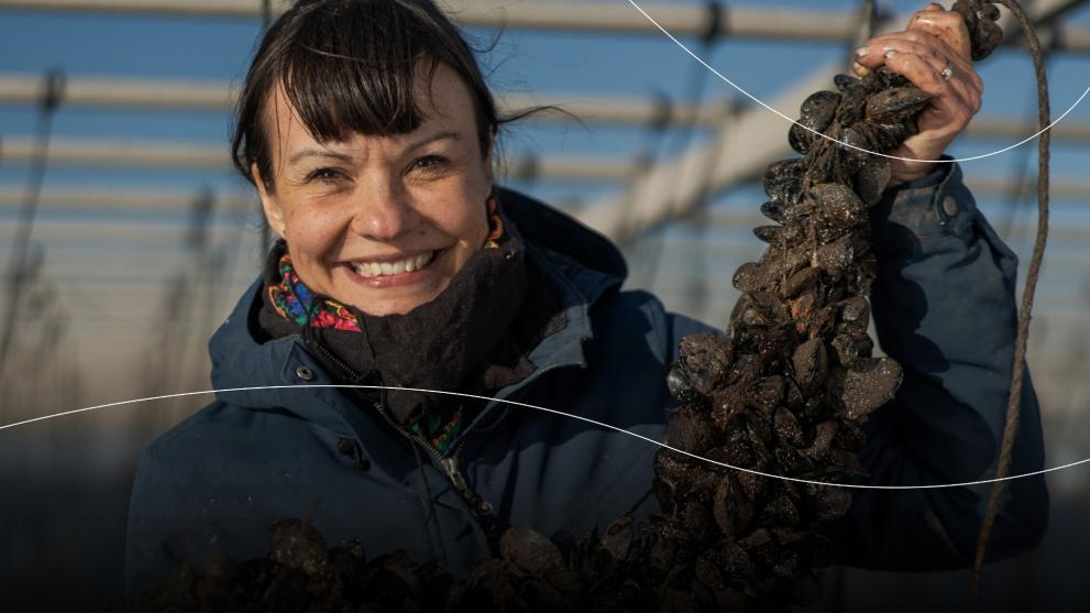 A smiling woman holds up mussels clustered on a rope.