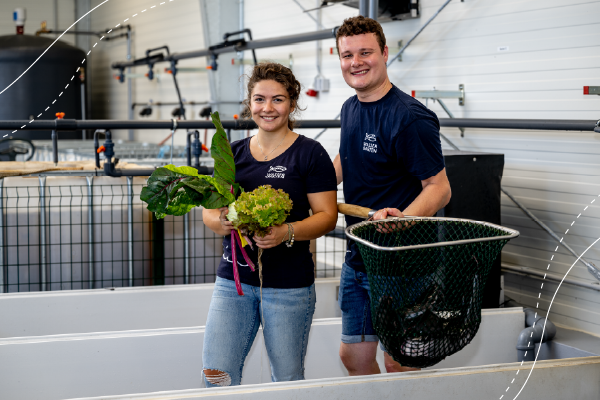A young woman and a young man stand in a warehouse. The woman is holding green vegetables, and the man is holding a landing net.
